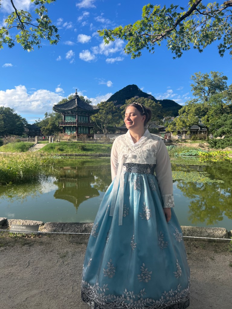 Woman in traditional Korean Hanbok informt of a lake and temple at Gyeongbokgung temple in Seoul, South Korea